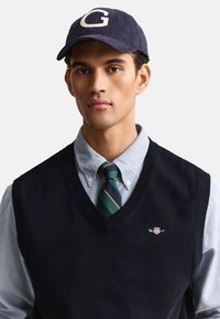 Giovane uomo che indossa un berretto blu navy con la lettera "G", camicia azzurro chiaro, cravatta a righe verde e blu e maglione nero senza maniche con piccolo logo.