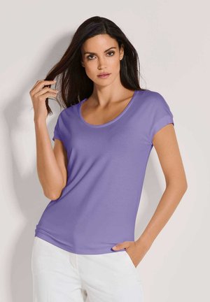 Lila Kurzarm-T-Shirt mit Rundhalsausschnitt, glatter Textur, enganliegendem Design, kombiniert mit weißen Hosen. Hände in den Taschen, Haare offen.