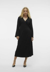 Zwarte wikkeljurk met lange mouwen en een gestructureerd oppervlak. Bevat een v-hals, een strik in de taille en valt tot halverwege de kuit. Gecombineerd met zwarte hakken.