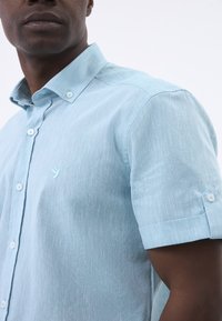 Hellblaues kurzärmeliges Hemd mit Knopfleiste aus strukturiertem Stoff, mit einem spitzen Kragen und einem kleinen Vogel-Logo auf der Brust.