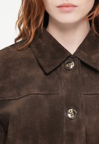 Nærbillede af en person med rødt hår iført en mørkebrun ruskindsjakke med store knapper og bred krave.