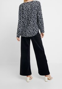 Žena v námořnické květinové blůze s dlouhým rukávem, černých volných kalhotách a bílých sandálech na podpatku, stojící na pozadí čisté bílé.