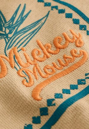Testo "Mickey Mouse" ricamato in arancione su tessuto beige con bordo geometrico blu e motivo floreale sul lato sinistro.
