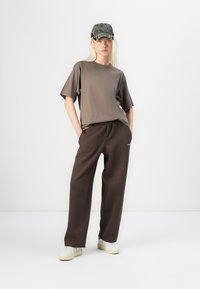 Bruine katoenen T-shirt met korte mouwen, gecombineerd met bruine joggingbroek, witte sneakers en een groene pet met contrasterende rand.