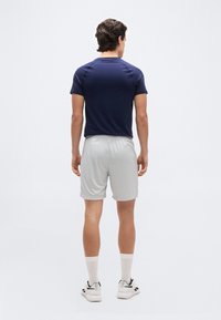 Ανδρας που στέκεται με την πλάτη προς τα εμπρός, φορώντας μπλε μπλούζα με κοντά μανίκια, ανοιχτό γκρι σορτς, λευκές κάλτσες και λευκά αθλητικά παπούτσια σε απλό φόντο.