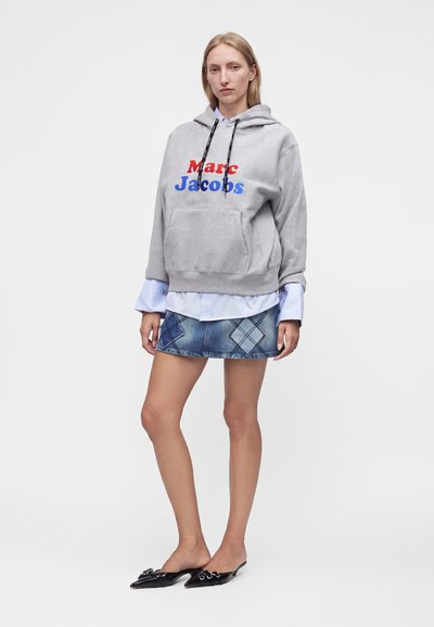 Γυναίκα φοράει γκρι φούτερ Marc Jacobs, λευκό πουκάμισο από κάτω, τζιν μίνι φούστα με διαμαντένια σχέδια και μαύρα μυτερά παπούτσια.