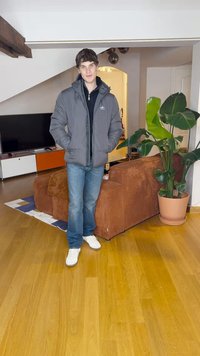 Pilkas pūkuotas švarkas uždedamas ant juodos marškinėlių, mėlynų džinsų ir balta sportinių batų. Fone yra rudas kordūrovas sofa ir vazoninis augalas.