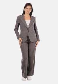 Blazer grigio in suede su misura e pantaloni a gamba larga abbinati, con una maglietta aderente color crema sotto. Presenta una chiusura con un bottone sul blazer.