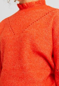 Detailní pohled na osobu oblečenou v jasně oranžovém svetru s vysokým výstřihem, který má texturované detaily ve tvaru V.