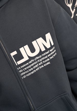 Sweat à capuche zippé gris foncé avec un grand texte blanc "MUL" et un texte plus petit inversé en dessous sur la poitrine.