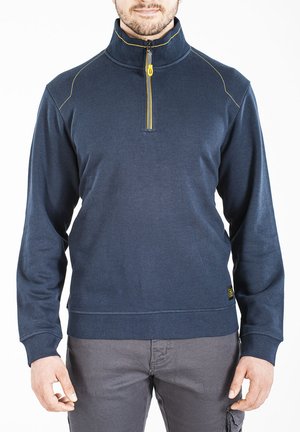 Homme portant un pull à fermeture éclair quart de zip bleu marine avec tirette jaune et pantalon gris, debout sur fond blanc.