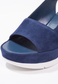 Sandale à talon en daim bleu marine avec un bout ouvert et une large bride. Dotée d'une semelle intérieure bleue lisse et d'une semelle extérieure en caoutchouc blanc.