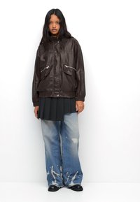Giacca bomber in finta pelle marrone con tasche con zip, gonna plissettata nera e jeans baggy azzurri con macchie di candeggina, indossati con scarpe nere.