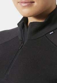 Detailní záběr osoby v černém pulovru se zapínáním na čtvrt zip, s vyvýšeným límcem a malým logem „HH“ na rameni.