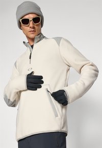 Fleece-pullover i kremfarge med grå detaljer, halv-glidelås foran og sidelomme med glidelås. Svart hansker og grå strikket lue fullfører antrekket.