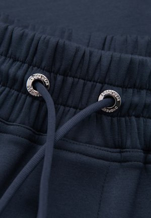 Elastic navyblåt taljebånd med snor og metaløjer indgraveret med "OMBREE WEAR" på mørkt stof.