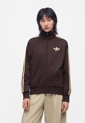 Jeune personne aux cheveux courts et noirs portant une veste marron foncé zippée avec des rayures beiges et un logo, et un pantalon beige ample.