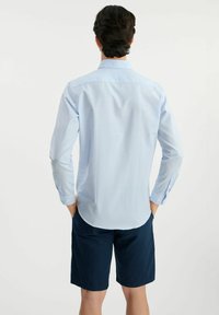 Jasnoniebieska koszula w kratę z długimi rękawami, casualowy krój i zaokrąglony dół, zestawiona z granatowymi szortami. Materiał wydaje się gładki i przewiewny.