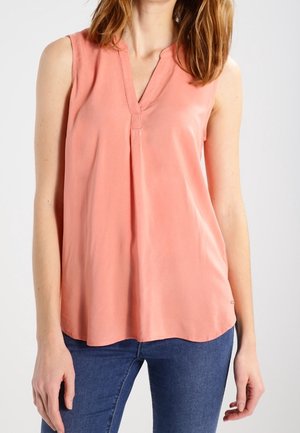 Ærmeløs koralrosa bluse med V-hals båret af en person, der står, kombineret med blå jeans mod en ensartet lys baggrund.