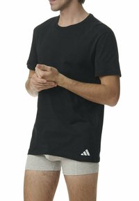 Mężczyzna w czarnej koszulce i jasnoszarych bokserkach, trzymający jedną rękę w drugiej przed białym tłem.