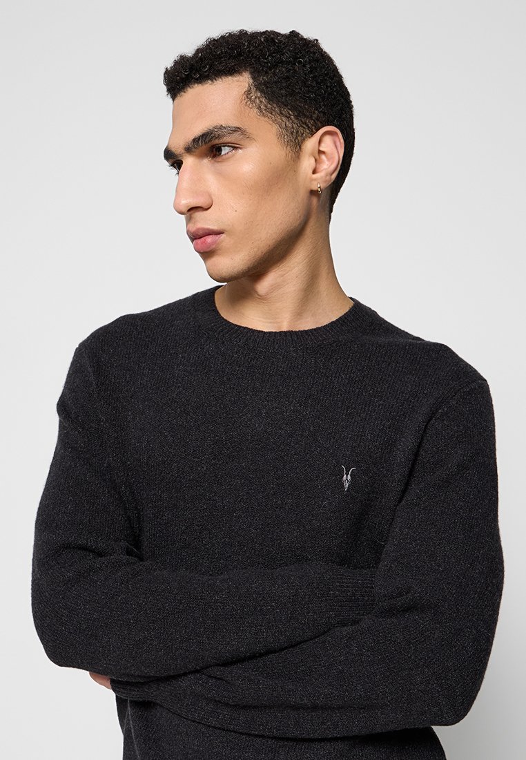 Junger Mann mit kurzen, lockigen schwarzen Haaren, der einen schwarzen Pullover mit einem kleinen gestickten Ziegenlogo trägt und zur Seite schaut, während er die Arme verschränkt.