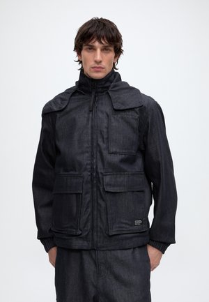Jeune homme aux cheveux foncés portant une veste noire zippée avec poches devant et un pantalon foncé assorti sur un fond clair uni.
