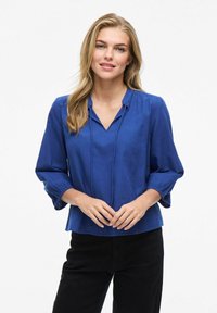 Blusa leggera blu con scollo a V e maniche a tre quarti. Presenta polsini elasticizzati e una vestibilità rilassata, abbinata a pantaloni neri.