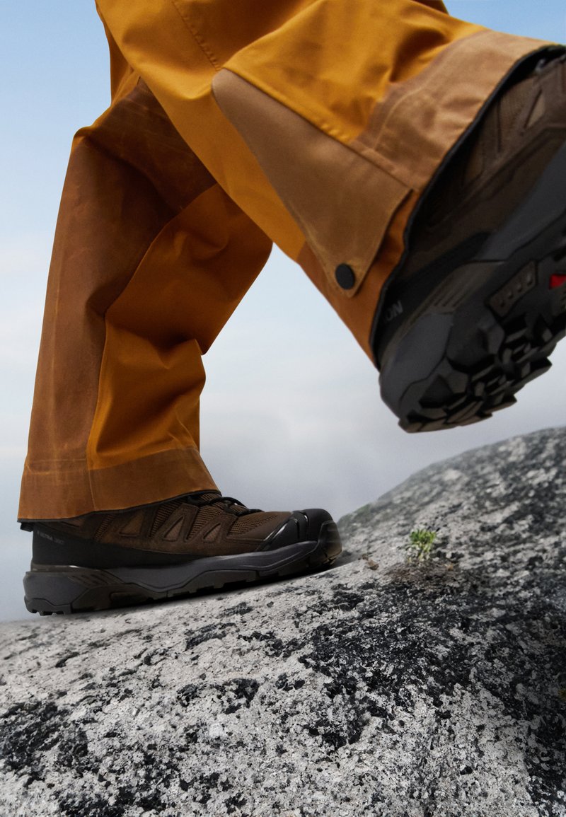 Brązowe buty trekkingowe z teksturowaną siateczką i gumową podeszwą, połączone z żółtymi wodoodpornymi spodniami, stojące na skalistym podłożu.