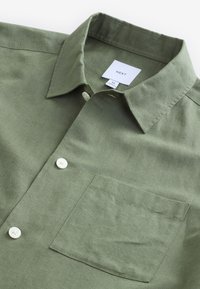 Olivengrøn skjorte med knapper foran, lavet af tekstureret stof, med spids krave, enkelt brystlomme og hvide knapper. Mærkets label er synlig.