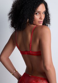 Soutien-gorge en dentelle rouge avec des bretelles ajustables fines, présentant un motif floral brodé. Le dos est équipé d'une fermeture à crochet et d'une texture lisse.