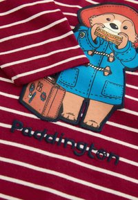 Chemise rayée rouge et blanche avec un motif de l'ours Paddington, portant un manteau bleu et tenant un sandwich, avec "Paddington" brodé en dessous.
