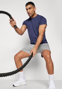 Jūras spēku īsām piedurknēm krekls un pelēki sporta šorti, ko valkā persona, kas izmanto melnu kaujas virkni, ar baltām sporta kurpēm un īsām zeķēm.