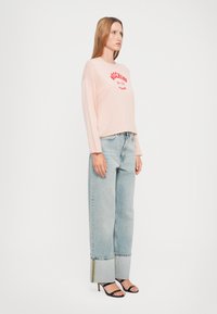 Chemise à manches longues rose clair avec du texte rouge "Moschino Jeans", assortie à un jean large bleu clair avec un accent jaune sur l'ourlet. Talons hauts noirs à lanières.