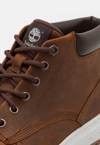Brun läderankelsko med vadderad krage, metallöglor, bruna snören och en vit gummisula med en strukturerad yta. Timberland-logga synlig.