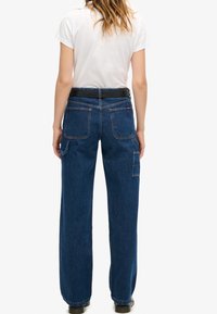 Vaqueros de mezclilla azul con un ajuste relajado, que presentan dos bolsillos traseros y un bolsillo lateral. Cinturón negro con un acabado suave. Camiseta blanca lisa.