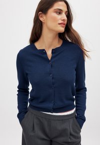 Marineblauer Cardigan aus Strickstoff, mit Rundhalsausschnitt, Knopfverschluss und gerippten Bündchen, getragen zu grauen, maßgeschneiderten Hosen.