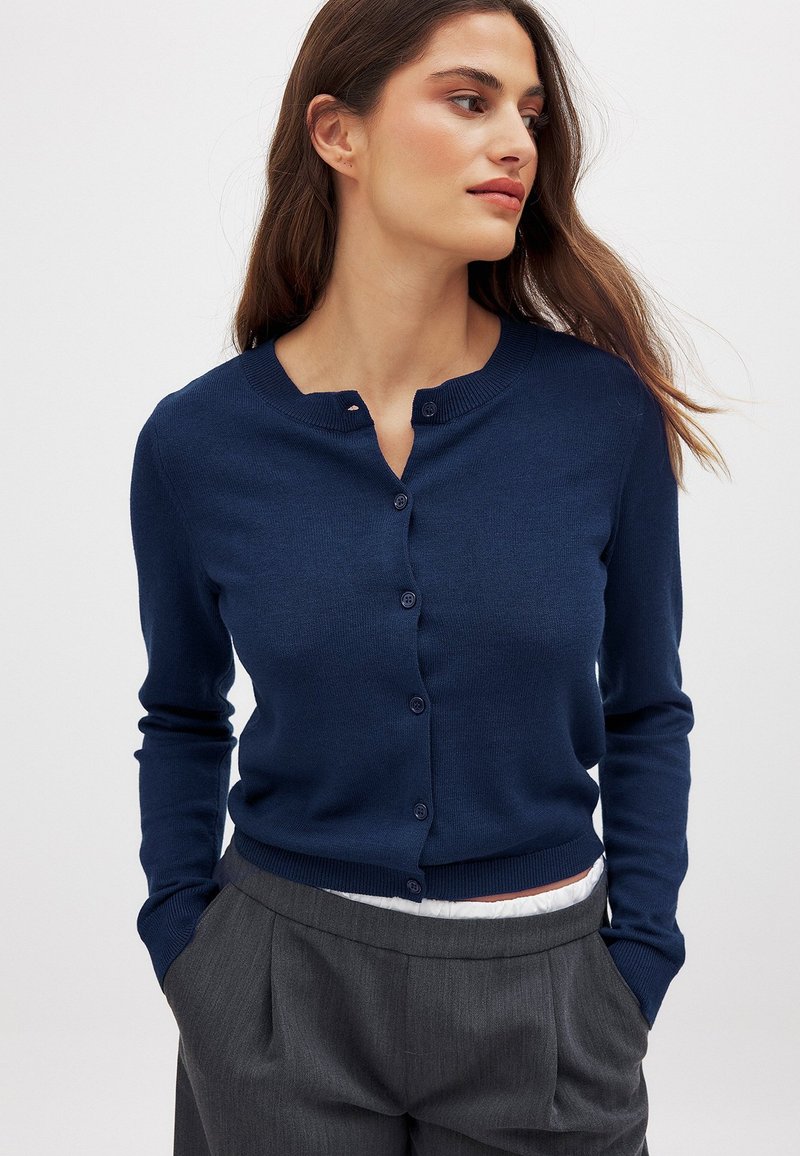 Marineblauer Cardigan aus Strickstoff, mit Rundhalsausschnitt, Knopfverschluss und gerippten Bündchen, getragen zu grauen, maßgeschneiderten Hosen.