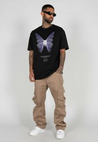 Sort grafisk t-shirt med et lilla sommerfuglemotiv, parret med lysebeige cargo-bukser og hvide sneakers. Solbriller fuldender looket.