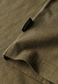 Tessuto di cotone verde oliva con una finitura testurizzata, caratterizzato da un'etichetta "SUPERDRY" blu cucita nella cucitura, che mette in mostra dettagli di cucitura pulita.