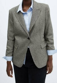 Černobílý sako vzorované v pepitku s rozparkovými klopami, zapínáním na jeden knoflík a dvěma předními kapsami, vrstvené přes světle modrou košili.