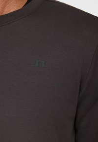 Mörkgrå långärmad tröja med ribbat mönster och rund halsringning. Har en liten svart logotyp på övre vänstra bröstet.