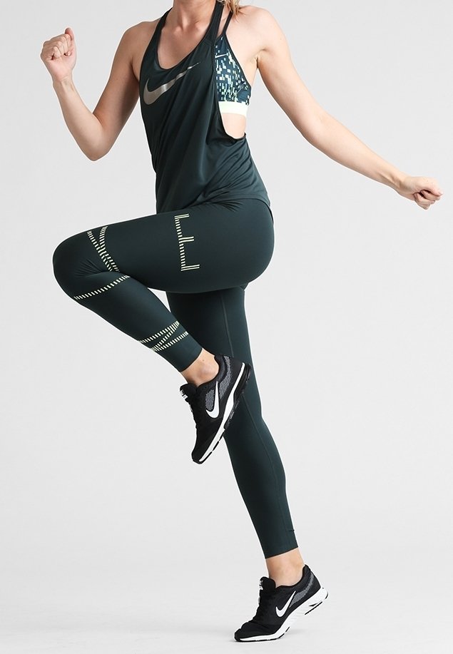 Donkergroene sportieve tanktop met uitsnijdingen en een geometrisch patroon, gecombineerd met bijpassende donkergroene legging met witte accenten en strepen. Zwarte hardloopschoenen maken de outfit af.