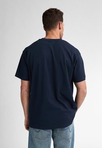 Námornícky modré bavlněné tričko s krátkým rukávem, kulatým výstřihem a volným střihem. Hladká textura, viditelné švy, kombinováno s světle modrými džíny.