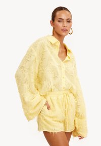 Gele, textuurrijke, oversized shirt met kraag, gecombineerd met bijpassende shorts met trekkoord. Beiden hebben een zachte, afgerafelde uitstraling.