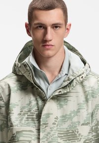 Ελαφρύ μπουφάν με ανοιχτό πράσινο καμουφλάζ. Διαθέτει φερμουάρ, κουκούλα και τσέπη στο στήθος με λεπτομέρεια κουμπιού.