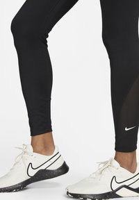 Nogi w czarnych legginsach i białych trampkach Nike z czarną podeszwą i sznurówkami na białym tle.