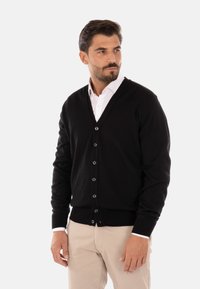 Cardigan nero a scollo a V realizzato in tessuto morbido, con chiusura a bottoni e polsini a costine. Indossato sopra una camicia bianca e pantaloni beige.