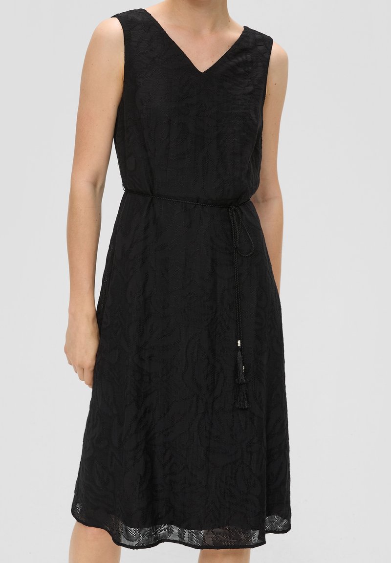 Robe noire sans manches longueur genou avec un motif subtil de feuilles, encolure en V et fine ceinture tressée avec des pompons, nouée à la taille.