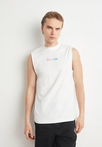 Camiseta sin mangas de algodón blanca con el texto "Esto es Amor" en colores del arcoíris, con un cuello redondo y corte relajado.