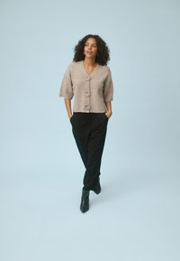Femme aux cheveux bouclés marchant avec assurance, portant un cardigan beige à fleurs boutonné, un pantalon noir et des bottines noires, sur un fond bleu pâle.